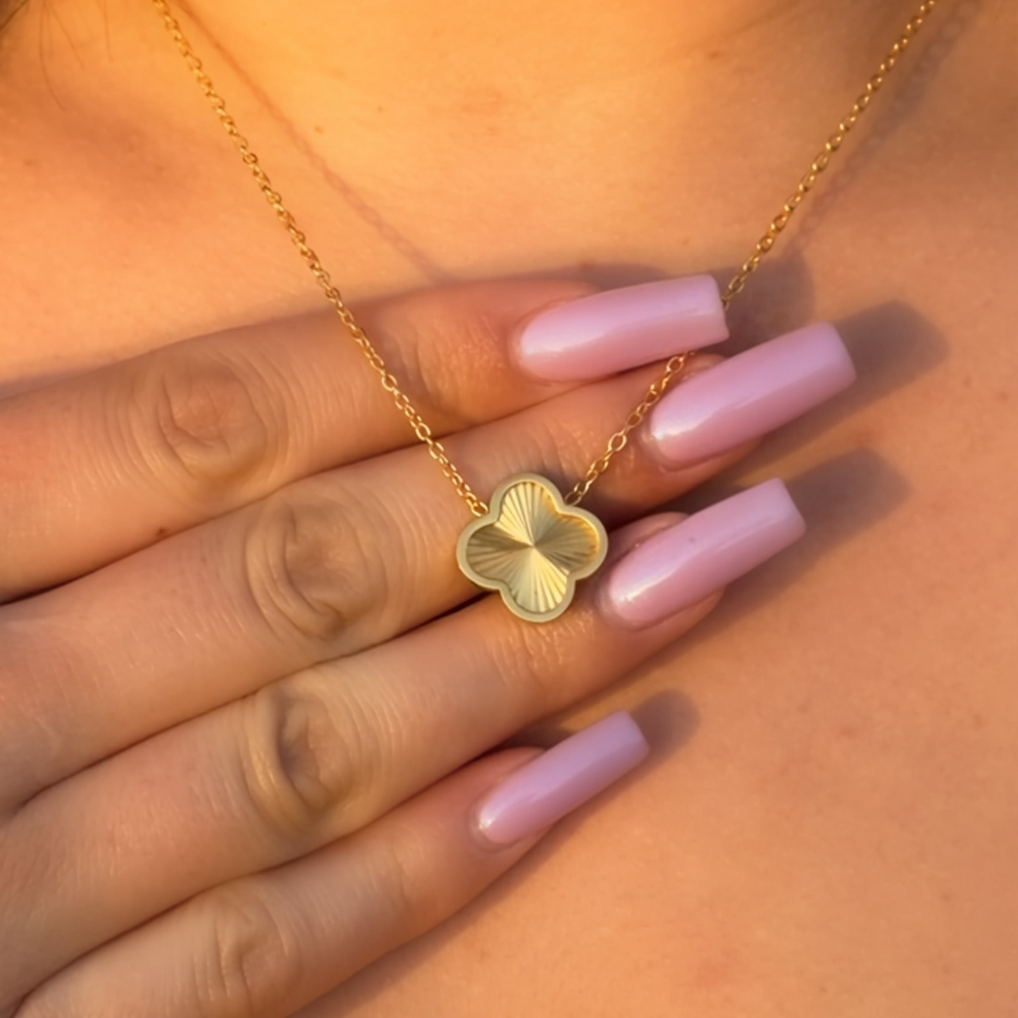 Gold necklace with a clover-shaped pendant worn by a person with pink nail polish.